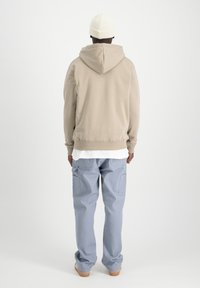 Sudadera beige con bolsillo tipo canguro, combinada con pantalones cargo azul claro que tienen bolsillos laterales. El modelo lleva un gorro blanco.