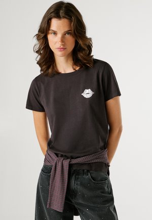 Jonge vrouw met schouderlang golvend haar draagt een zwarte t-shirt met een wit lippenlogo en een gestreepte trui om haar taille geknoopt, zwarte jeans met studs.