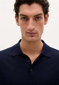 Marineblå polo-skjorte med klassisk krave, lavet af blødt stof, med tre mørke knapper og et diskret broderet logo på brystet.