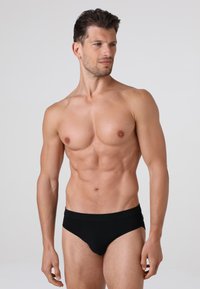 Slip bikini in cotone nero con una texture liscia, caratterizzati da una vita bassa e una vestibilità aderente, indossati da un modello maschile.