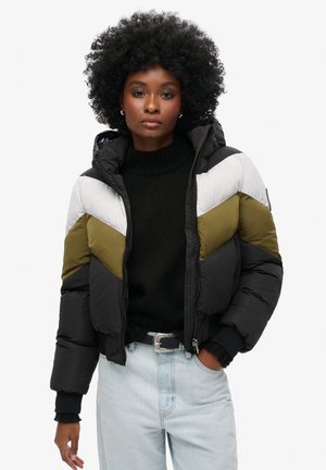 Puffjacke mit Kapuze, mit schwarzen, olivgrünen und weißen Zickzackmustern, Reißverschluss und gerippten Bündchen. Getragen mit einem schwarzen Pullover.