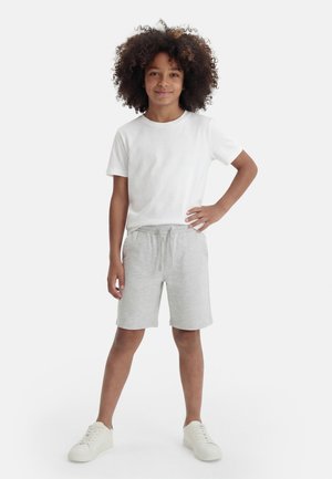 Copil cu păr creț stă cu o mână pe șold, purtând tricou alb, pantaloni scurți gri deschis și adidași albi pe fundal simplu.