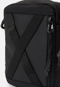 Borsa in tessuto nera con superficie texturizzata, cinturini incrociati, chiusura con zip e accessori opachi. Design compatto e rettangolare.