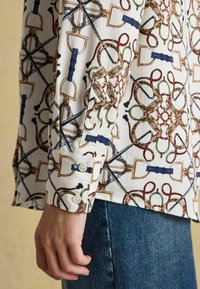 Sleeve of white long-sleeve shirt with equestrian print featuring stirrups, ropes, and buckles, worn with blue denim jeans on a person's arm.