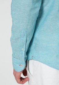 Gros plan d'une personne portant une chemise bleu clair à manches longues et un pantalon blanc, montrant les détails du poignet de la chemise et de l'ourlet latéral.