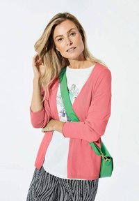 Korallefarbener Cardigan aus leichtem Material, mit V-Ausschnitt, getragen über einem weißen T-Shirt mit Druckgrafik, kombiniert mit gemusterten schwarzen Hosen und einer grünen Umhängetasche.