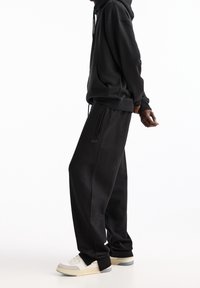 Schwarzer Hoodie mit Kordelzug und frontaler Tasche, kombiniert mit lockeren schwarzen Sweatpants. Das Outfit hat ein dezentes Logo-Detail.