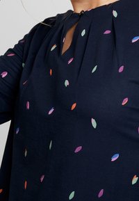 Tyrkysová blůza s detailními záhyby, zdobená barevnými vzory listů v zelené, růžové, oranžové a modré barvě; akcent v podobě klíčové dírky u výstřihu.