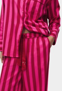 Pasiasty komplecik piżamy w fuksjowym i różowym kolorze, składający się z koszuli na guziki oraz spodni na elastycznej talii. Koszula posiada kieszonkę na piersi; spodnie mają detal w postaci przewiązania.