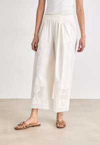 Pantalons blancs à jambes larges en tissu léger avec des motifs brodés et des découpes au niveau de l'ourlet. Associés à des sandales à glisser marron.