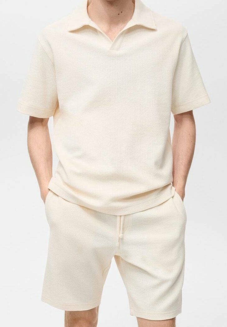 Homme portant un polo en maille couleur crème à manches courtes avec col et un short en maille assorti, mains dans les poches, sur fond blanc.