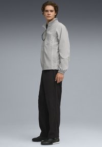 Ljusgrå zip-up jacka med vita accenter, elastiska ärmslut och ståkrage, tillsammans med svarta byxor och svarta sneakers.