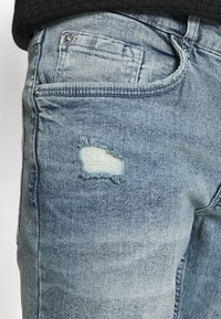 Jeans in denim blu chiaro con strappo effetto consumato. Design classico a cinque tasche, cuciture visibili e finitura del tessuto strutturata.