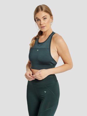 Ung kvinde med flettet hår iført mørkegrøn ærmeløs crop top og høj talje leggings, stående mod en ensartet lys baggrund.