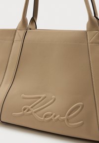 Sac à main en cuir beige avec deux poignées et le nom "Karl" embossé en cursive sur le panneau avant.