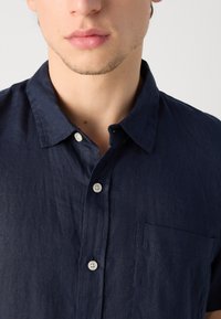 Navyblauw linnen shirt met een button-down kraag, een voorzak en witte knopen. De stof heeft een licht gestructureerd uiterlijk.