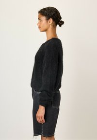 Juodas purvinas megztinis su ilgomis rankovėmis, laisvo kirpimo, dėvimas su tamsiai mėlyna džinsine sijonine, kuri turi nelygų kraštą, matomas iš šono.