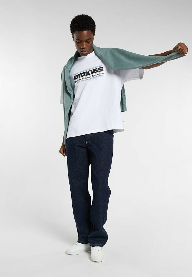 Young man wearing a white Dickies T-shirt, dark jeans, white sneakers, with a green sweater draped over his shoulders.