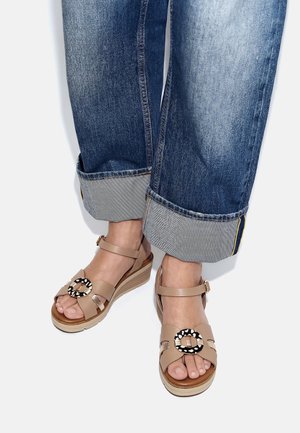 Füße in beigen Keilsandalen mit rundem Schnallen mit Tiermuster und aufgekrempelten blauen Jeans auf weißem Hintergrund.