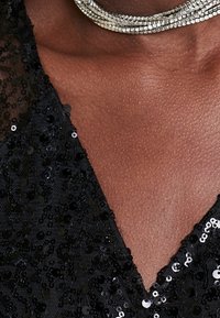 Un tissu en sequins noirs forme un décolleté en V profond, accentué par un collier en argent à plusieurs brins avec des maillons rectangulaires, mettant en valeur la texture et l'éclat.