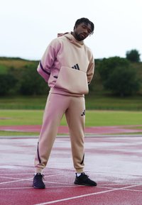 Moletom atlético bege com um bolso frontal e detalhes em roxo, combinado com calças jogger beigues correspondentes com riscas pretas, usadas com ténis pretos.