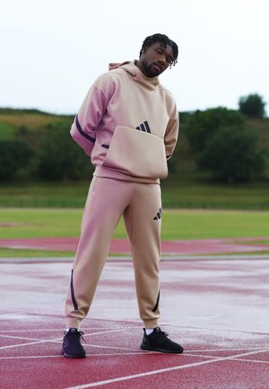 Beige athletic hoodie with a front pocket and purple accents, paired with matching beige joggers featuring black stripes, worn with black sneakers.