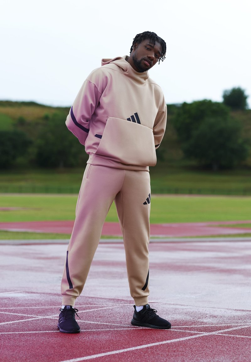 Moletom atlético bege com um bolso frontal e detalhes em roxo, combinado com calças jogger beigues correspondentes com riscas pretas, usadas com ténis pretos.