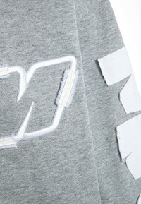 Tejido de sudadera gris con bordados y parches en relieve en blanco. Presenta una textura suave, un diseño de logo distintivo y acentos bien cortados.