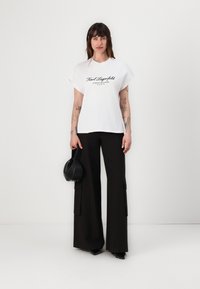 White cotton t-shirt featuring a black printed logo, paired with high-waisted black wide-leg trousers with side pockets and slits.