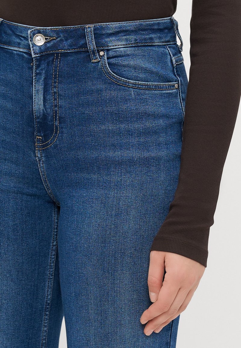 Jeans in denim blu a vita alta con una finitura liscia, dotati di chiusura frontale con bottone e design a cinque tasche. Top aderente marrone scuro parzialmente visibile.