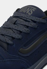 Gros plan d'une basket Vans bleu marine avec des lacets texturés et une bande latérale noire sur une semelle en caoutchouc sombre.