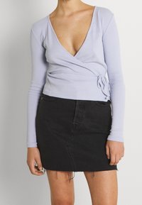 Kvinde iført en lyse lilla langærmet indpakningsbluse og en sort denim miniskirt med to franske kanter mod en ensfarvet baggrund.