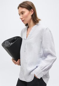 Rövid barna hajú nő fehér blúzban, egy nagy fekete bőr táskát tart egy egyszerű világos háttér előtt.