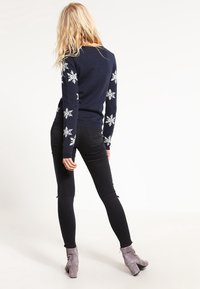 Modell vänder sig bort, iklädd en marinblå tröja med vita snöflingemönster på ärmarna och svarta byxor, kompletterat med grå ankelstövlar.