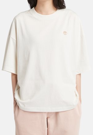 Frau trägt ein lockeres weißes T-Shirt mit kurzen Ärmeln und einem kleinen braunen Baumlogo auf der linken Brust sowie hellrosa Hosen.
