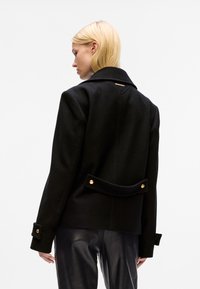 Zwarte wollen jas met grote kraag, achterkantdetails en accenten van goudkleurig beslag, met een relaxte pasvorm en gestructureerde mouwen.