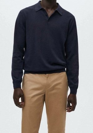 Personne portant un polo à manches longues bleu marine, rentré dans un pantalon beige, debout devant un fond clair uni.