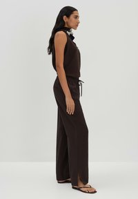 Bruine mouwloze top en bijpassende wijde broek met trekkoord in de taille en zijsplitten. Model draagt zwarte slippers op een neutrale achtergrond.
