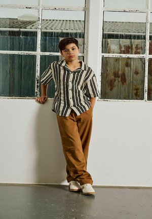 Jongen met kort donker haar leunt op een witte vensterbank, draagt een gestreept overhemd met korte mouwen en knopen, een bruine broek en witte sneakers.