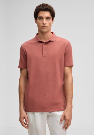 Jonge man die een polo met korte mouwen in stoffig roze draagt en een witte broek, tegen een eenvoudige lichte achtergrond.