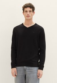 Pulover negru cu decolteu în V, realizat dintr-un material tricotat texturat, cu mâneci lungi și un design ajustat, asortat cu blugi gri deschis.