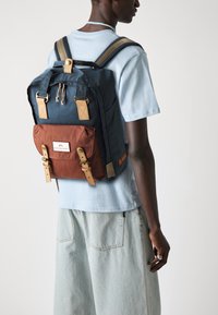 Navy and brown backpack with leather accents, top handle, adjustable straps, and a front pocket secured by buckles and a logo patch.