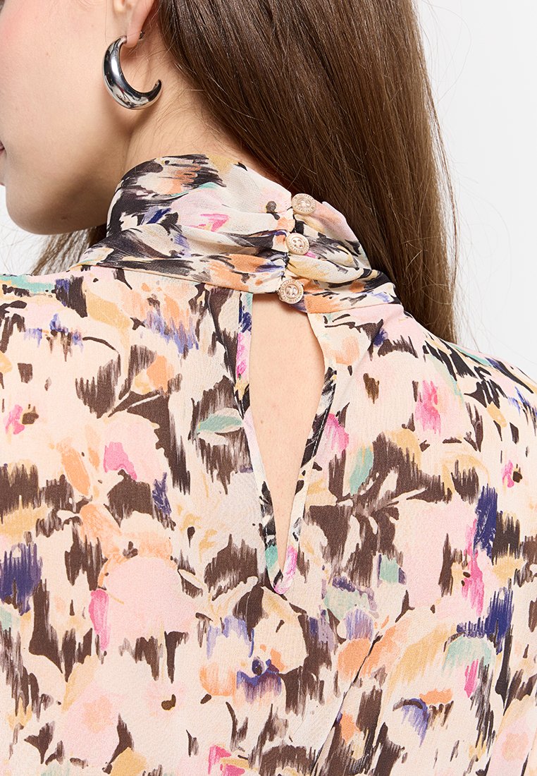 Kwiatowa bluzka z wysokim kołnierzem, miękkiego materiału, w pastelowych kolorach, z otworem w tylnej części i zapięciami na guziki przy kołnierzu.