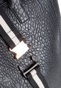 Svart syntetisk ryggsäck med strukturerad yta, justerbar svart och vit randig rem, samt metalliska detaljer med glänsande gul accent.