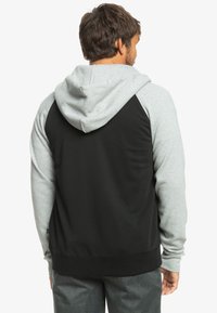 Pánska mikina s kapucňou so sivým predným dielom a rukávmi, čiernym chrbtom, ribkovaným lemom, hladkou textúrou a klokanou vreckom.