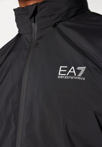 Črna, lahka jakna z visokim ovratnikom in sprednjim zadrgo. Na levi prednji strani ima bele črke logo EA7 Emporio Armani.