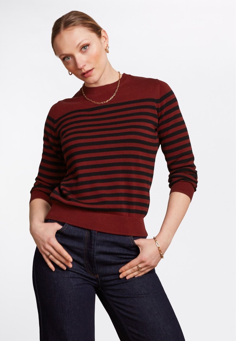 Maglione a strisce rosse e nere con scollo rotondo e polsini a costine. Orlo arrotondato, texture morbida, abbinato a jeans in denim scuro.