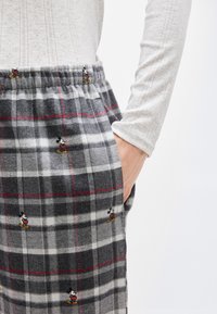 Personne portant un pantalon gris à carreaux avec de petites impressions de Mickey Mouse et un haut blanc texturé à manches longues, main reposant dans la poche.