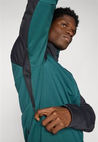Türkise und schwarze Sportjacke mit zippered Ventilation, dehnbarem Material, hohem Kragen und kontrastierenden Nähten. Das Modell hebt den Arm, um das Detail zu zeigen.