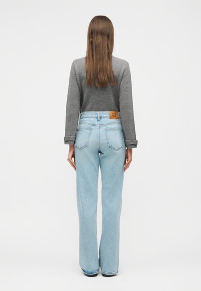 Γυναίκα που στέκεται με την πλάτη, φορώντας γκρι μακρυμάνικη μπλούζα και ανοιχτό μπλε ψηλόμεσο παντελόνι με φαρδιά πόδια, μπροστά σε απλό φόντο.